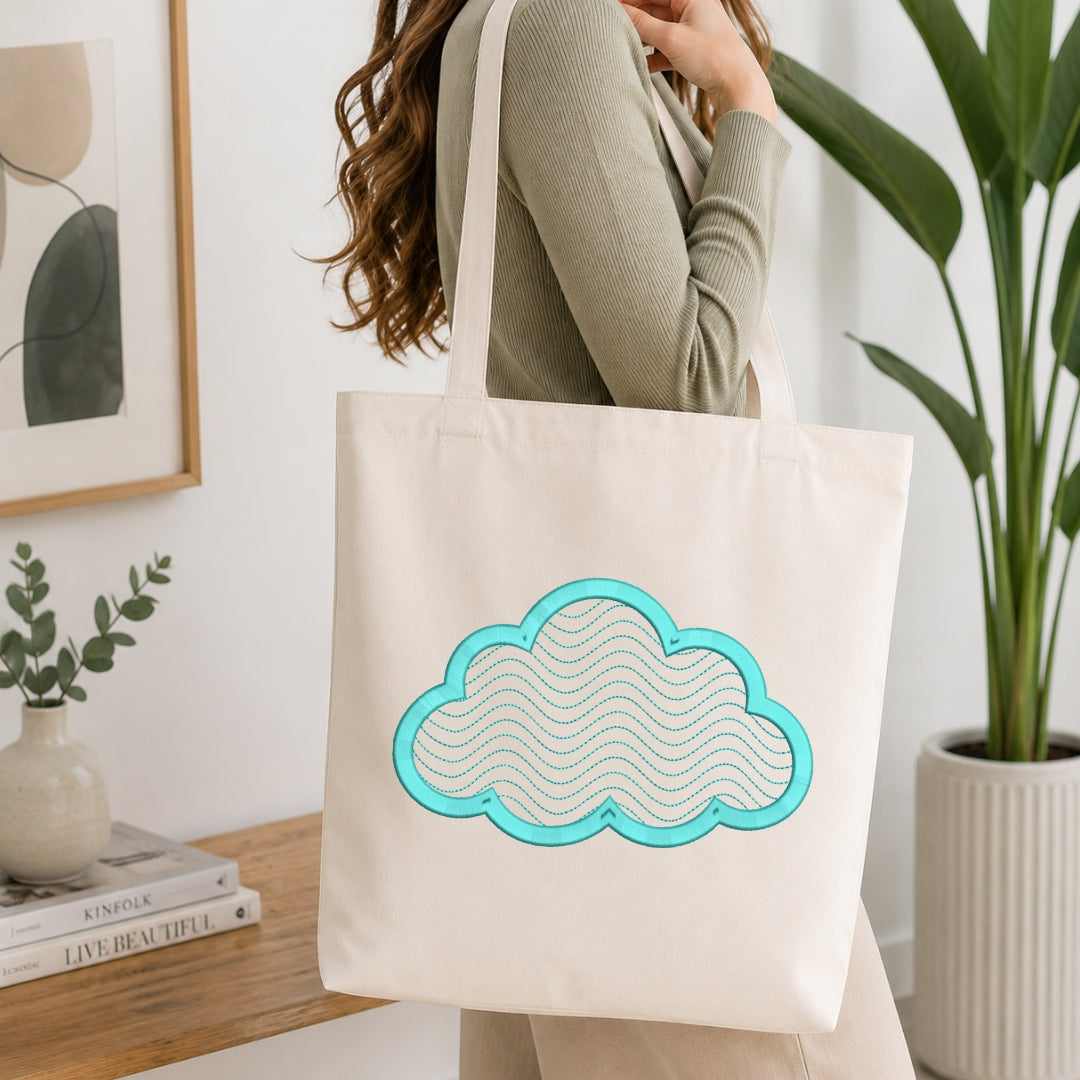 A person wearing a ribbed sage green top carries a cream canvas tote bag with a turquoise and white cloud design, set in a bright, modern room with a wooden table, books, and a potted plant.