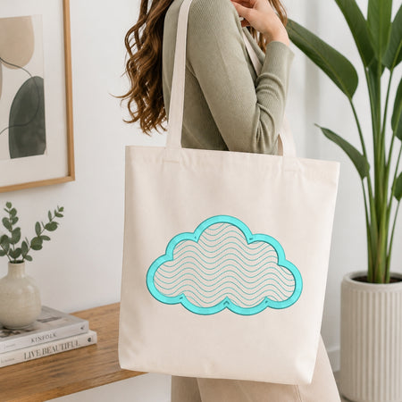 A person wearing a ribbed sage green top carries a cream canvas tote bag with a turquoise and white cloud design, set in a bright, modern room with a wooden table, books, and a potted plant.