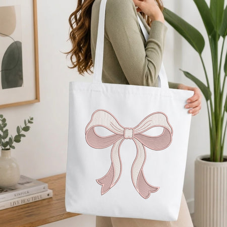 A woman in a sage green ribbed sweater carries a white canvas tote bag featuring a large pink and cream striped bow graphic on the front, set in a bright, modern room with a wooden table and potted plants.