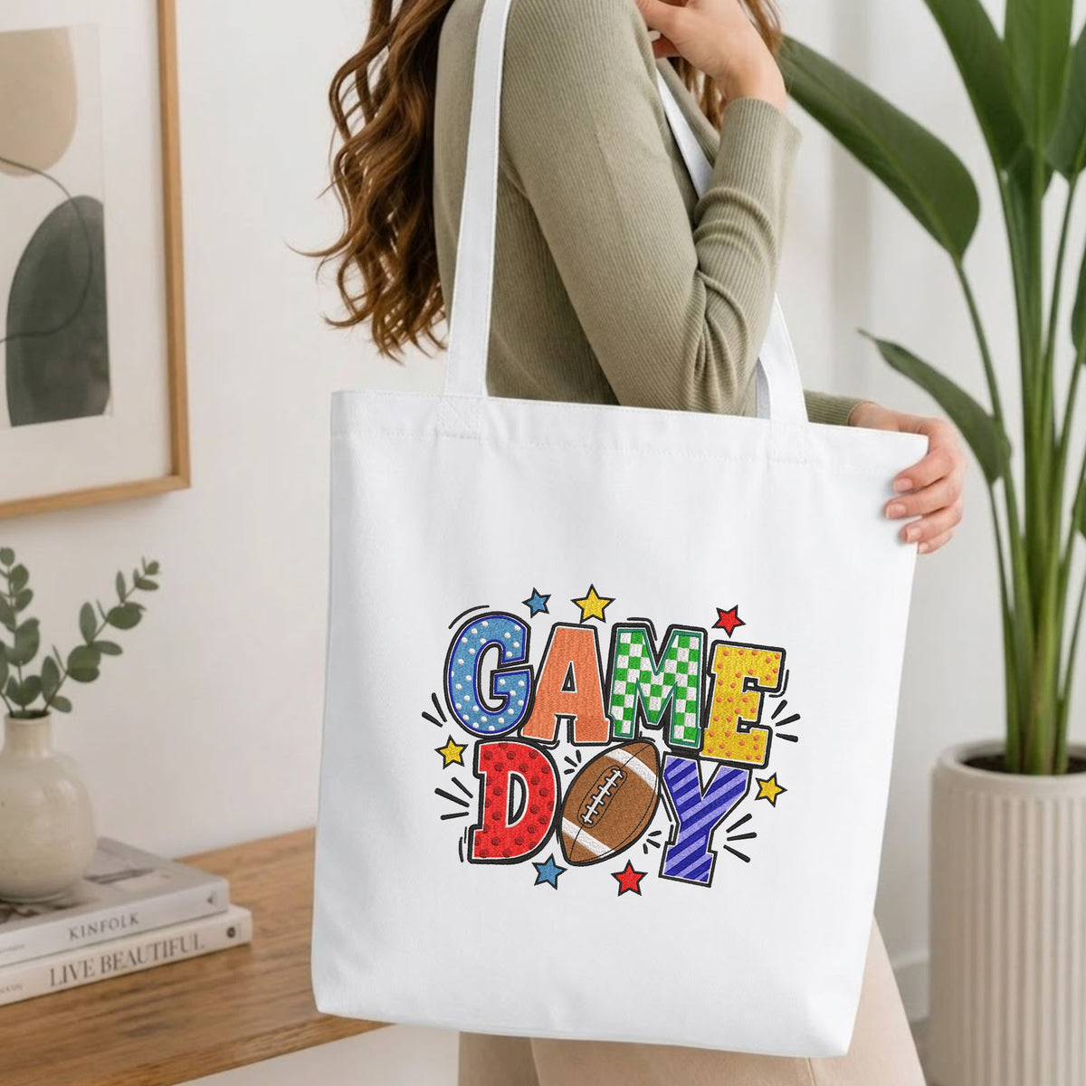 Woman carrying a white natural canvas tote bag featuring an embroidered "Game Day" football design in a bright, modern interior setting
