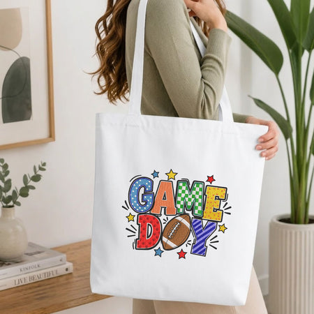 Woman carrying a white natural canvas tote bag featuring an embroidered "Game Day" football design in a bright, modern interior setting
