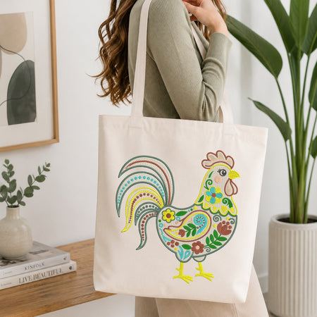 A woman wearing a sage green ribbed sweater carries a cream canvas tote bag decorated with a colorful, hand-drawn style rooster. The background is a bright, modern room with a wooden table, books, a small vase, and a large green potted plant.
