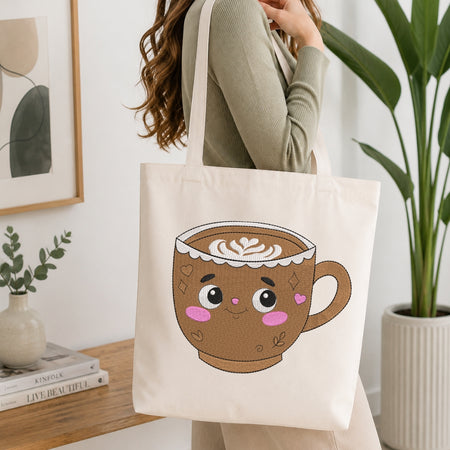 A person wearing a sage green ribbed long-sleeve top carries a cream canvas tote bag with a cute cartoon hot cocoa cup graphic on the front, set in a bright, modern room with a wooden table, books, and a potted plant.