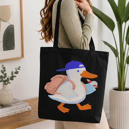 A person wearing a beige ribbed sweater carries a black canvas tote bag with a colorful embroidered duck design. The duck has a blue cap, pink backpack, white body, and orange beak and feet.