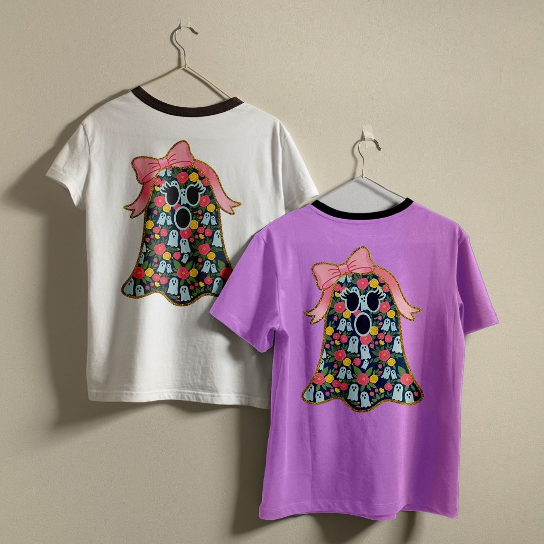 Two children's short-sleeve T-shirts hanging on hangers against a plain beige wall. The left shirt is white with a dark collar and features a large ghost graphic on the back in pink, purple, black, and multicolor.