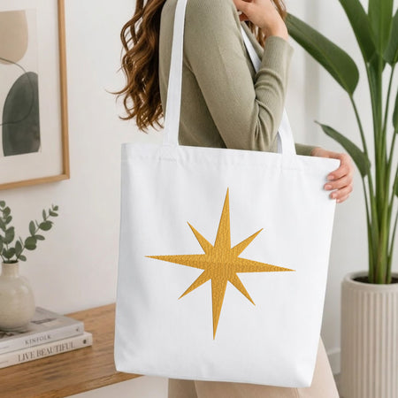 Woman carrying a white natural canvas tote bag featuring an embroidered golden starburst design in a bright, modern interior setting