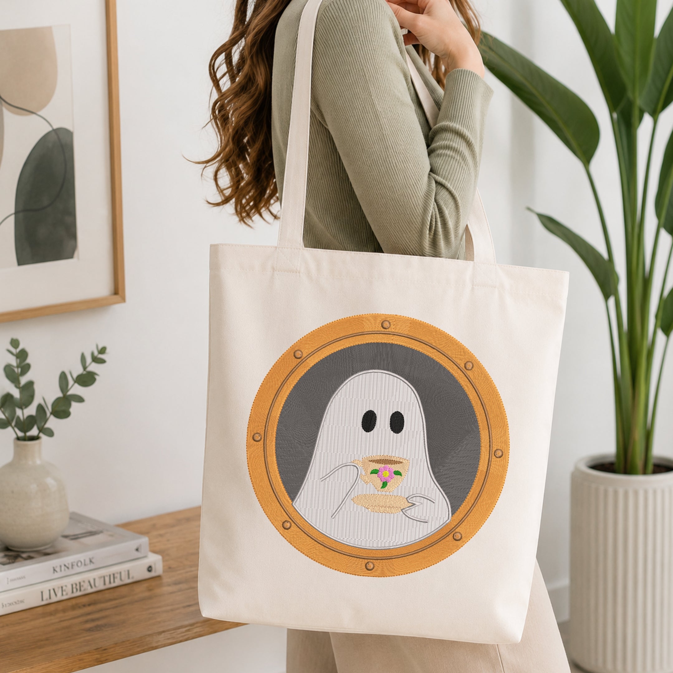 A woman wearing a sage green ribbed long-sleeve top carries a cream canvas tote bag with a graphic of a ghost peeking over a plate of pizza inside a porthole-style circle.