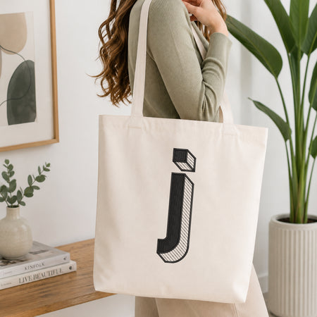 A woman wearing a sage green ribbed long-sleeve top carries a cream canvas tote bag with a large black stylized letter 'j' printed on the front, set in a bright, modern room with a wooden table, framed art, and a potted plant.