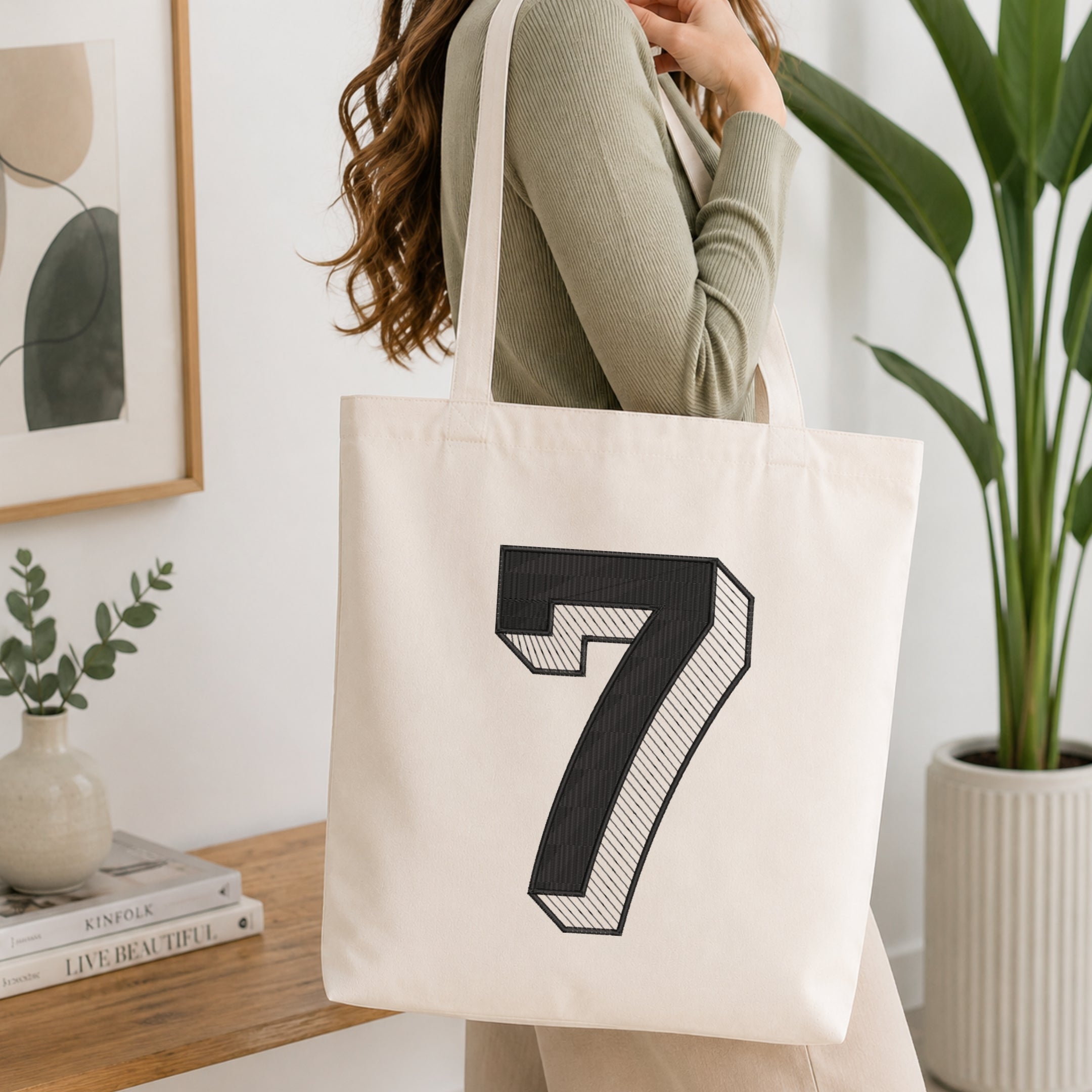 A woman wearing a sage green ribbed long-sleeve top carries a cream canvas tote bag with a large black 3D-style number 7 printed on the front, set in a bright, modern room with a wooden table, framed art, and a potted plant.