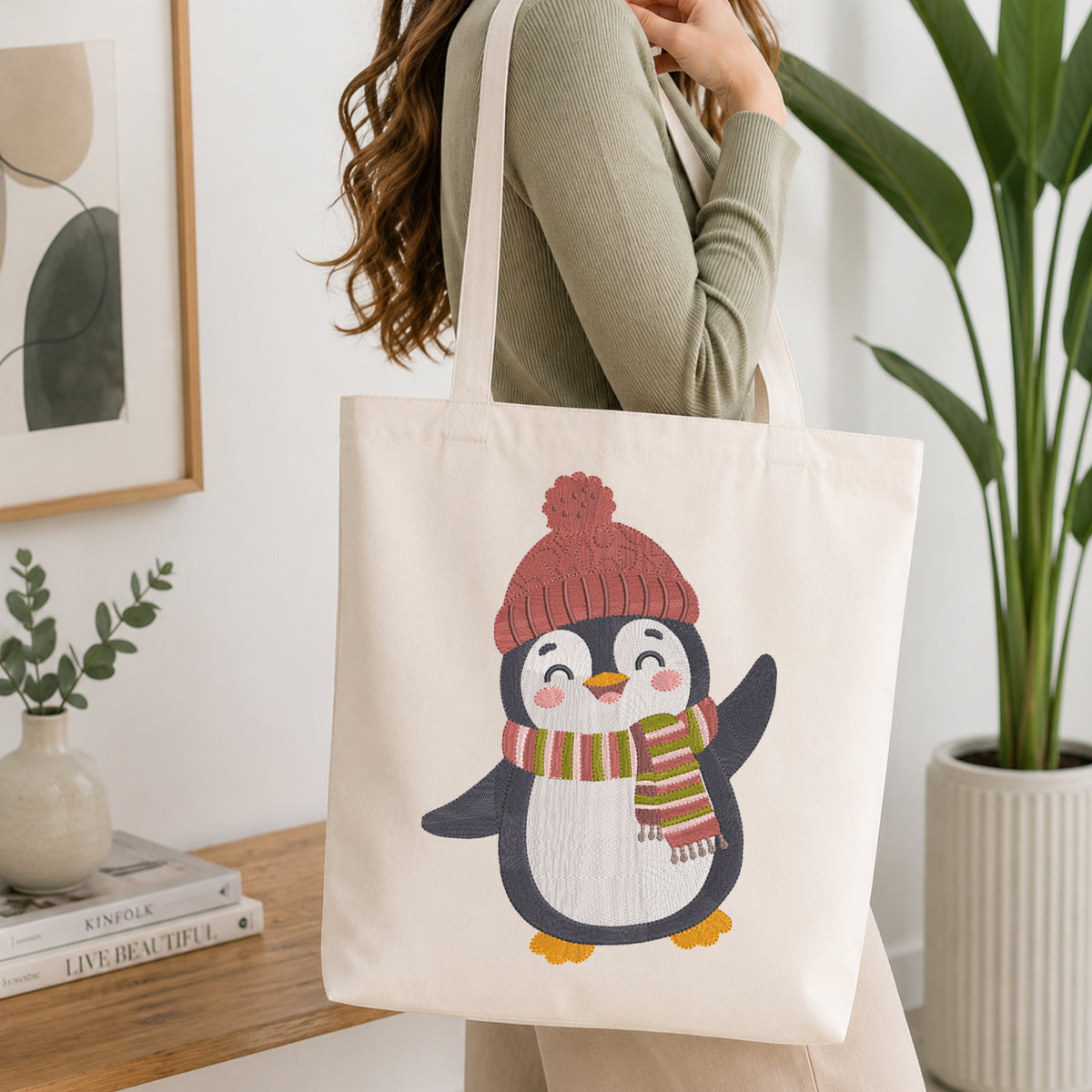 A woman wearing a sage green ribbed sweater carries a cream canvas tote bag featuring a cute penguin illustration wearing a pink knit hat and a striped green, orange, and pink scarf. The scene is set in a bright, modern room with a wooden side table, a small potted plant, and a large green plant in the background.