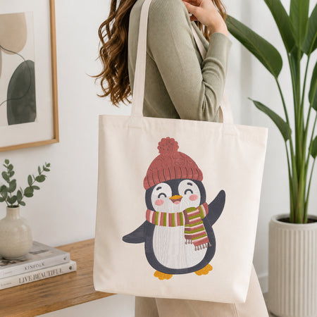 A woman wearing a sage green ribbed sweater carries a cream canvas tote bag featuring a cute penguin illustration wearing a pink knit hat and a striped green, orange, and pink scarf. The scene is set in a bright, modern room with a wooden side table, a small potted plant, and a large green plant in the background.
