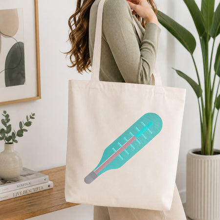 A woman wearing a sage green ribbed long-sleeve top carries a cream canvas tote bag with a stylized graphic of a teal thermometer on the front, set in a bright, modern room with a wooden table, books, and a potted plant.