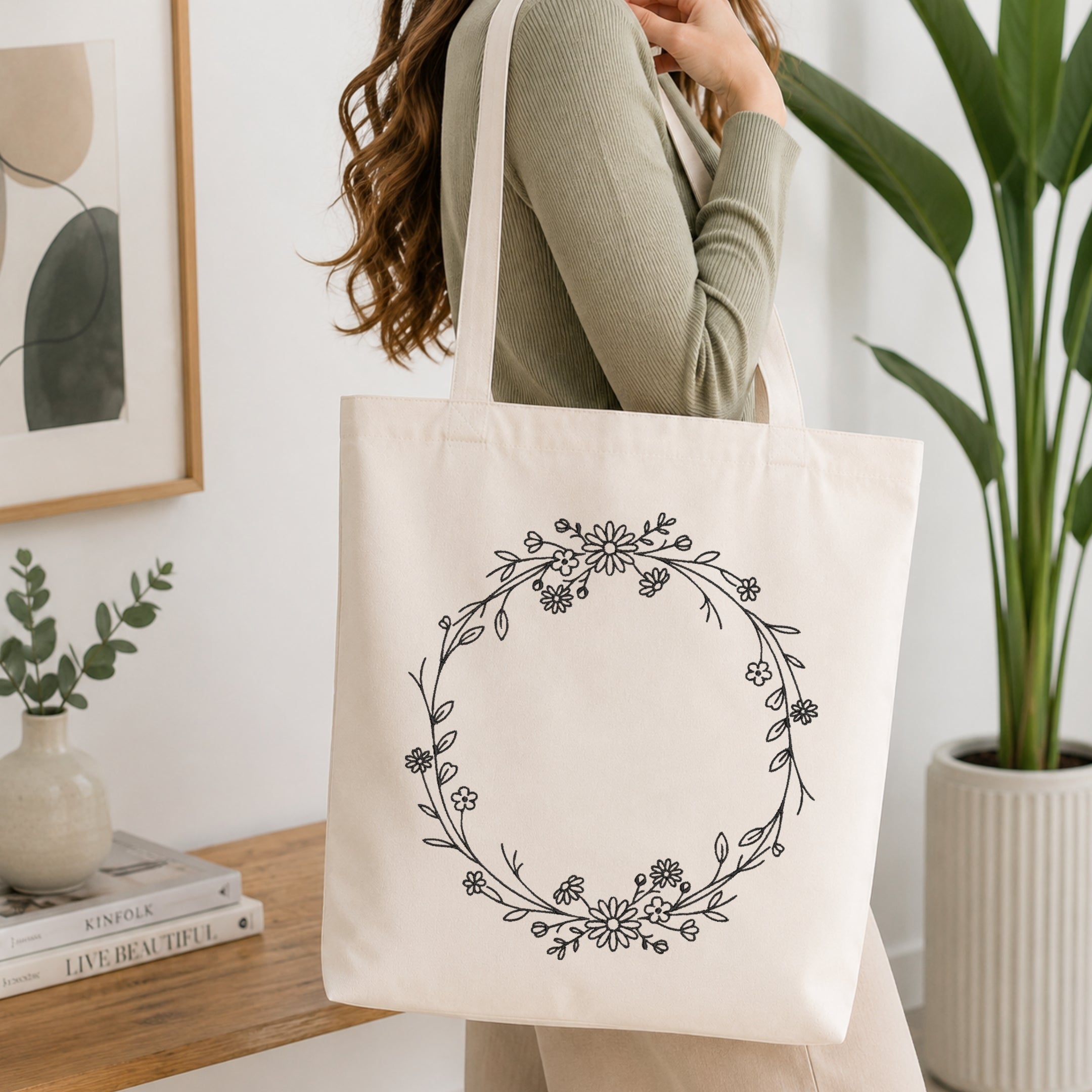 A person wearing a sage green ribbed sweater carries a cream canvas tote bag with a black floral wreath print, set in a bright, modern room with a wooden table, books, and a potted plant.