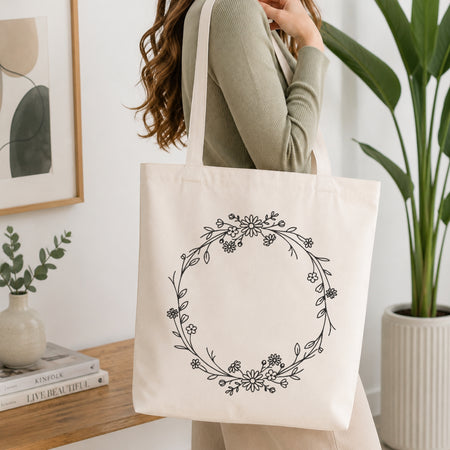 A person wearing a sage green ribbed sweater carries a cream canvas tote bag with a black floral wreath print, set in a bright, modern room with a wooden table, books, and a potted plant.