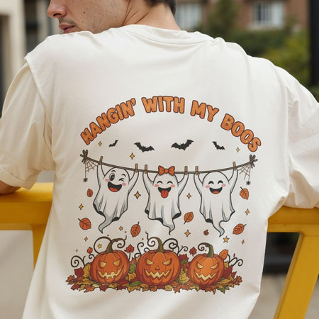 A person wearing a white graphic T-shirt, shown from the back. The shirt features a Halloween design with the text 'HANGIN' WITH MY BOOS' above three cartoon ghosts hanging on a clothesline, with pumpkins and autumn leaves below.
