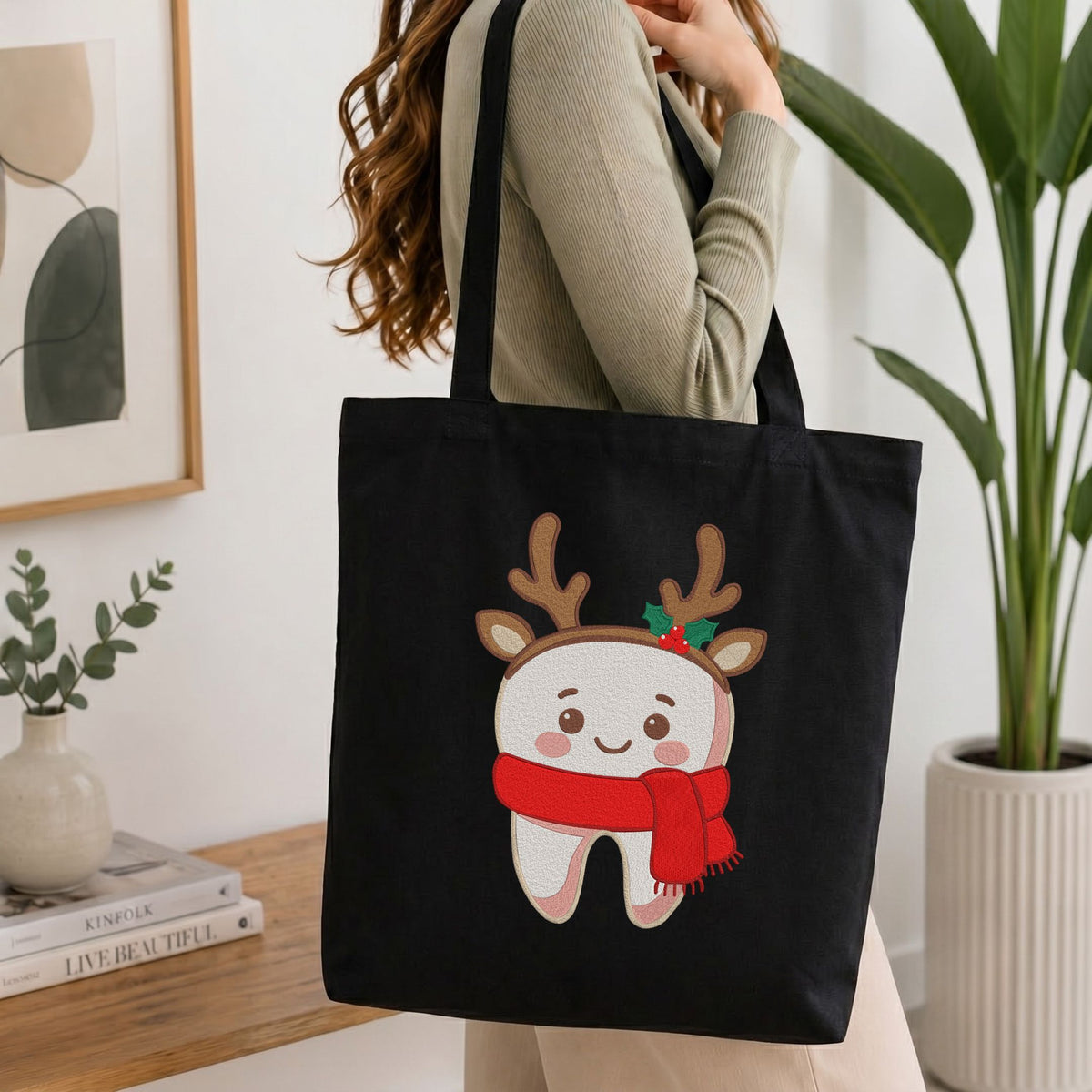 Woman carrying a black canvas tote bag featuring a cute smiling tooth wearing reindeer antlers and a cozy red scarf Christmas dental machine embroidery design in a bright interior setting