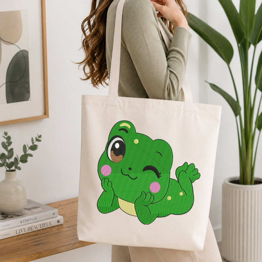A person wearing a ribbed sage green top carries a cream canvas tote bag featuring a cute winking green cartoon frog. The setting is a bright, modern room with a wooden table, books, a small potted plant, and a large green houseplant in the background.