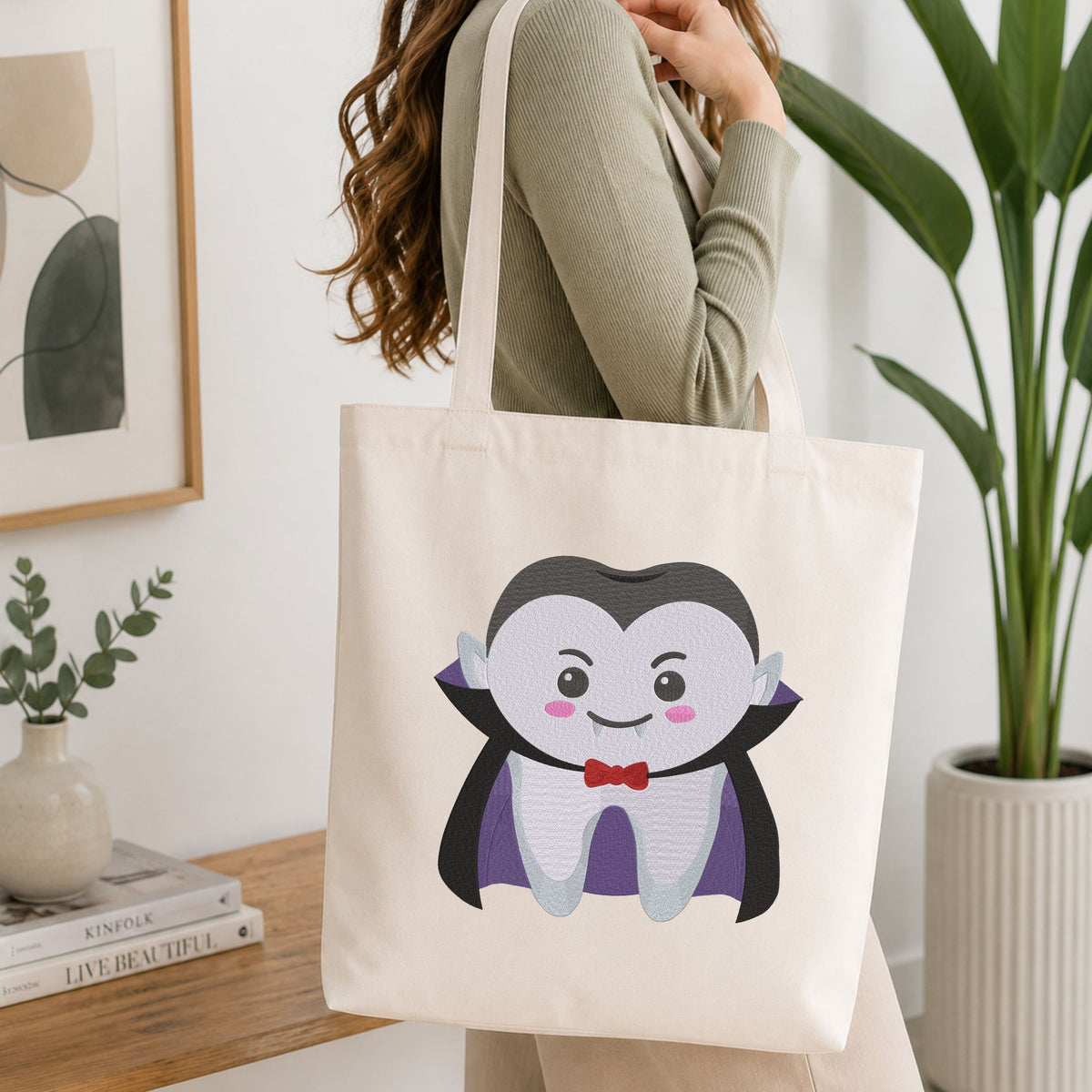 Woman carrying a white natural canvas tote bag featuring a cute vampire tooth Halloween dental machine embroidery design in a bright, modern interior setting