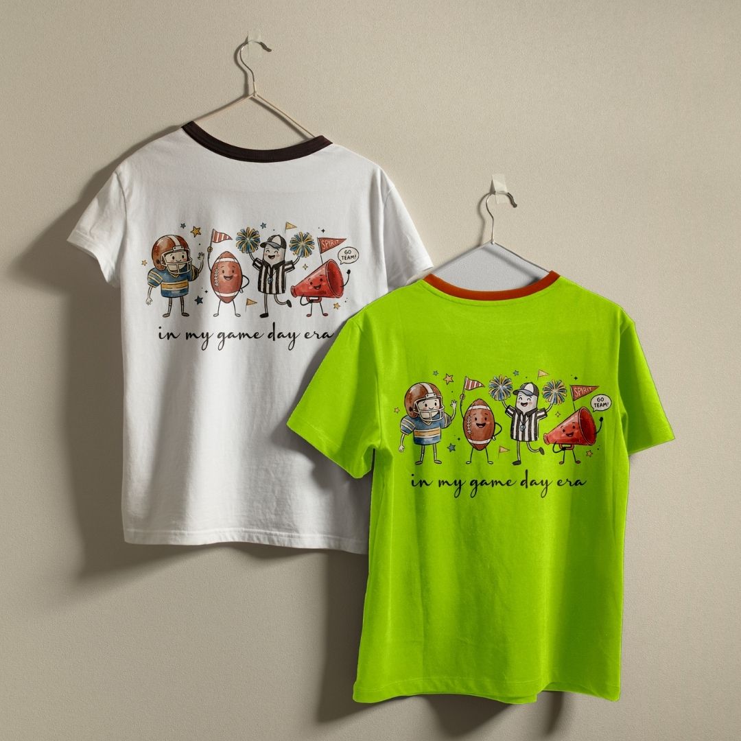 Two graphic t-shirts hanging on metal hangers against a plain beige wall. The left shirt is white with a dark brown collar and features a colorful cartoon graphic of sports figures and watermelon slices with the text 'in my game day cra'.