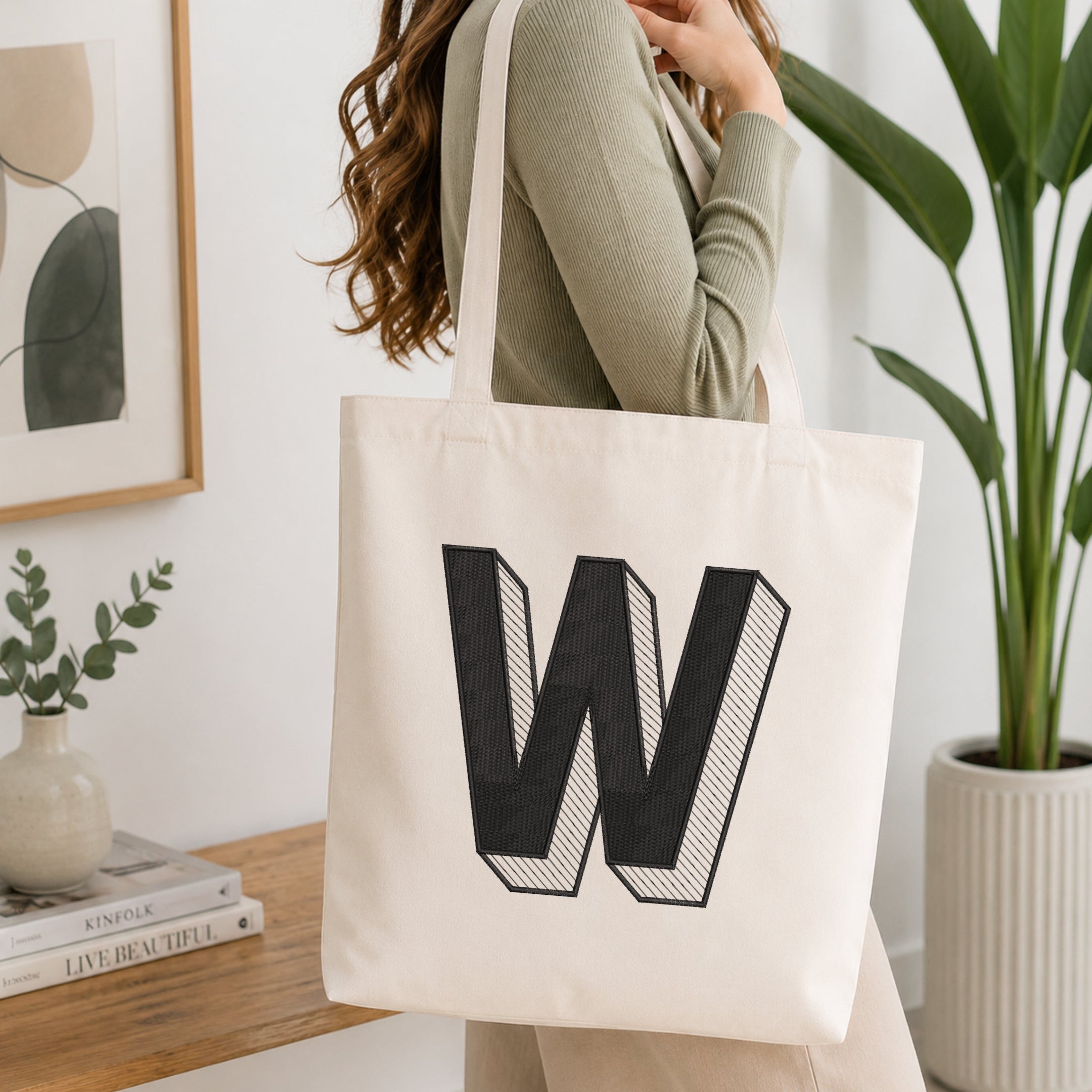 A person holding a natural canvas tote bag printed with an embroidered 3D block letter "W" design, set against a bright interior room background with green plants.