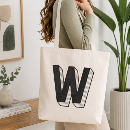A person holding a natural canvas tote bag printed with an embroidered 3D block letter "W" design, set against a bright interior room background with green plants.