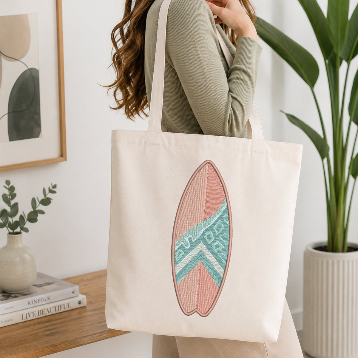 A woman wearing a sage green ribbed long-sleeve top carries a cream canvas tote bag with a pink and teal surfboard graphic on the front, set in a bright, modern room with a wooden table, framed art, and a potted plant.