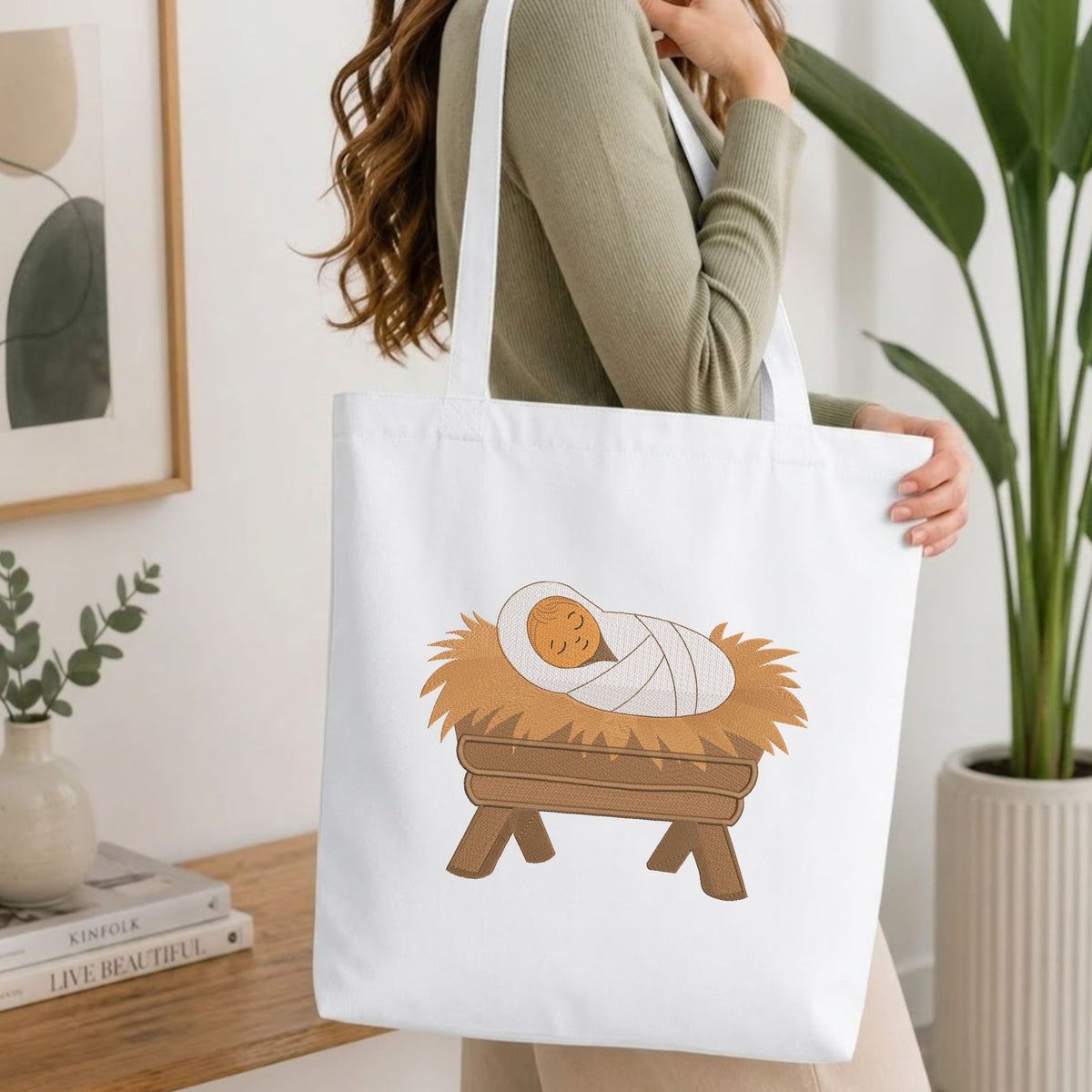 Woman carrying a white natural canvas tote bag featuring an embroidered baby Jesus in a manger design in a bright, modern interior setting.

