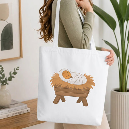 Woman carrying a white natural canvas tote bag featuring an embroidered baby Jesus in a manger design in a bright, modern interior setting.

