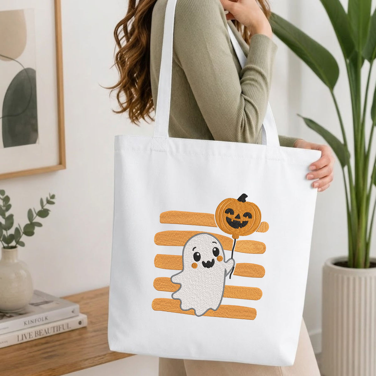Woman carrying a white natural canvas tote bag featuring an embroidered cute ghost design in a bright, modern interior setting