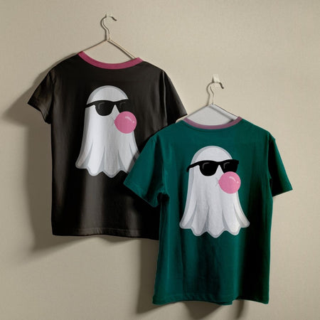 Two short-sleeve ringer t-shirts hanging on hangers against a plain beige wall. The left shirt is black with a pink collar and features a white ghost graphic wearing black sunglasses and blowing a pink bubble.