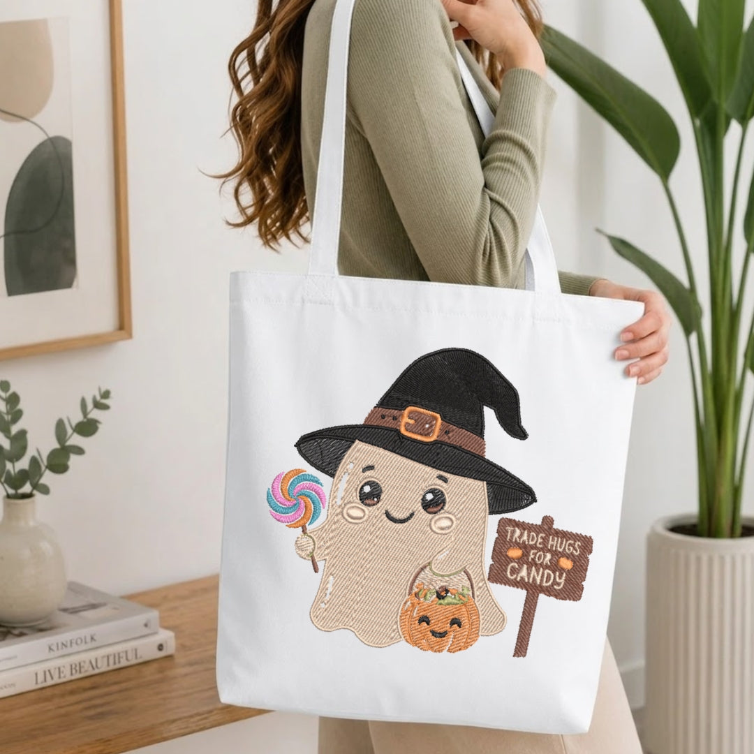 A woman holds a white canvas tote bag featuring a cute cartoon ghost wearing a black witch hat and holding a pumpkin and candy, with a sign that reads 'TRADE HUGS FOR CANDY'. The scene is set in a bright, modern room with a wooden table, books, and a potted plant.