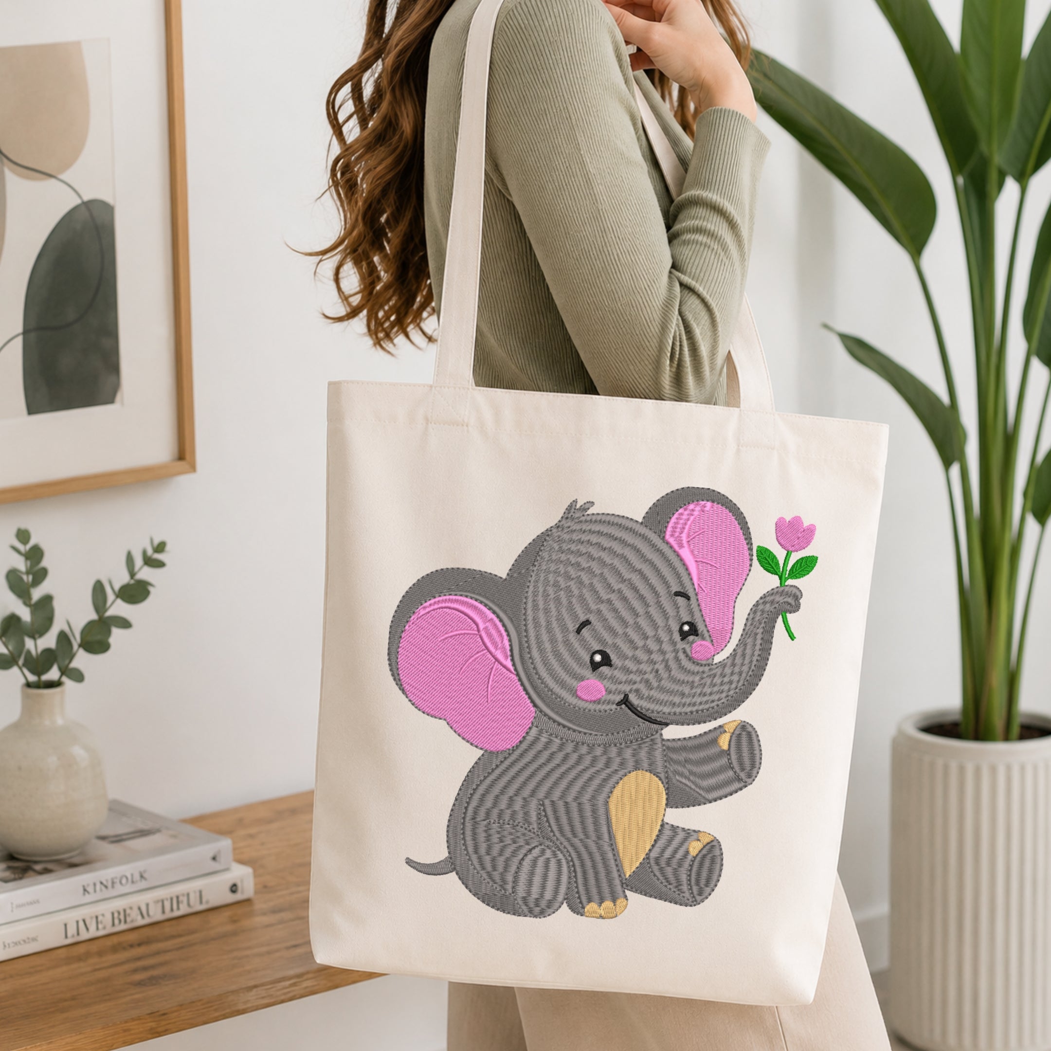 A woman wearing a sage green ribbed sweater carries a cream canvas tote bag with a large embroidered cartoon elephant holding a pink flower. The scene is set in a bright, modern room with a wooden table, books, a framed print, and a potted plant.