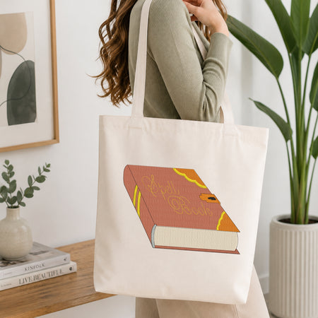 Woman carrying a natural canvas tote bag featuring an embroidered magical spell book design in a bright, modern interior setting.