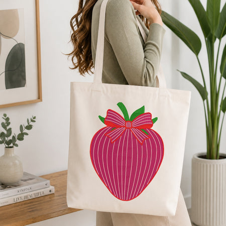 A woman wearing a sage green ribbed sweater carries a cream canvas tote bag with a large pink striped strawberry graphic featuring green leaves and a red bow.