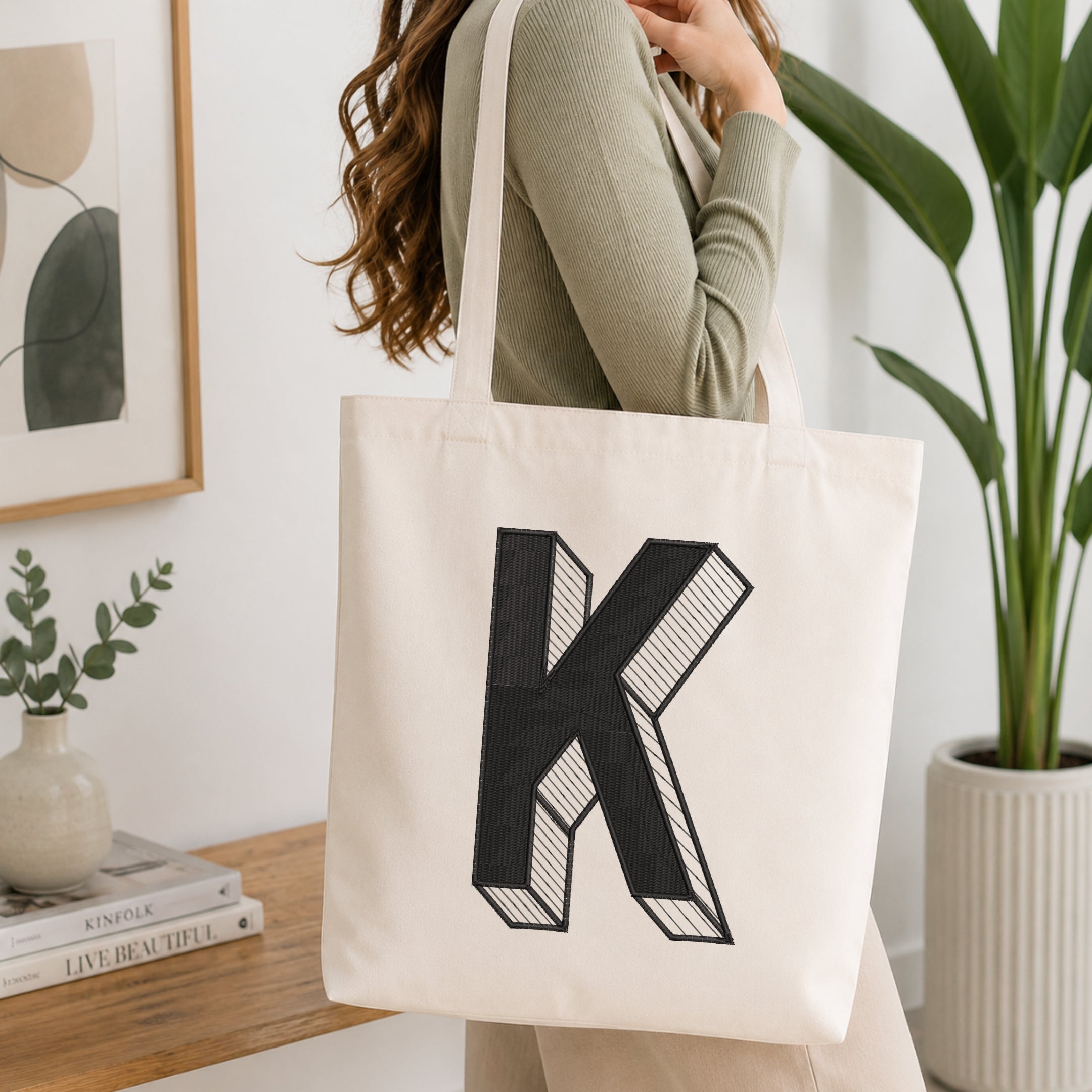 A woman wearing a sage green ribbed long-sleeve top carries a cream canvas tote bag with a large black block letter 'K' printed on the front, set in a bright, modern room with a wooden table, books, and a potted plant.