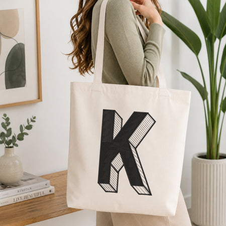 A woman wearing a sage green ribbed long-sleeve top carries a cream canvas tote bag with a large black block letter 'K' printed on the front, set in a bright, modern room with a wooden table, books, and a potted plant.