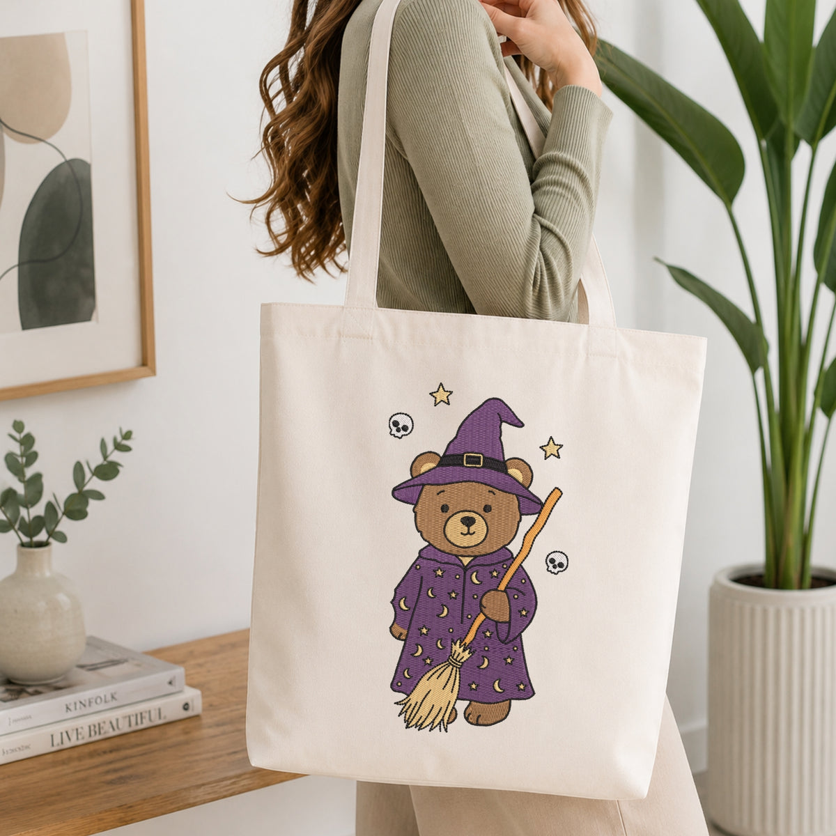 A woman wearing a sage green ribbed sweater carries a cream canvas tote bag with a cute illustration of a teddy bear dressed as a witch in a purple robe and hat, holding a broomstick. The background is a bright, modern room with a wooden table, books, a framed print, and a potted plant.