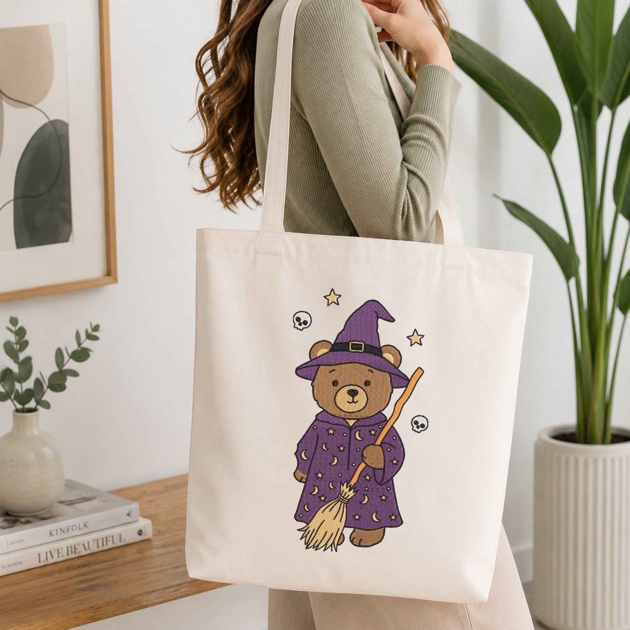 A woman wearing a sage green ribbed sweater carries a cream canvas tote bag with a cute illustration of a teddy bear dressed as a witch in a purple robe and hat, holding a broomstick. The background is a bright, modern room with a wooden table, books, a framed print, and a potted plant.