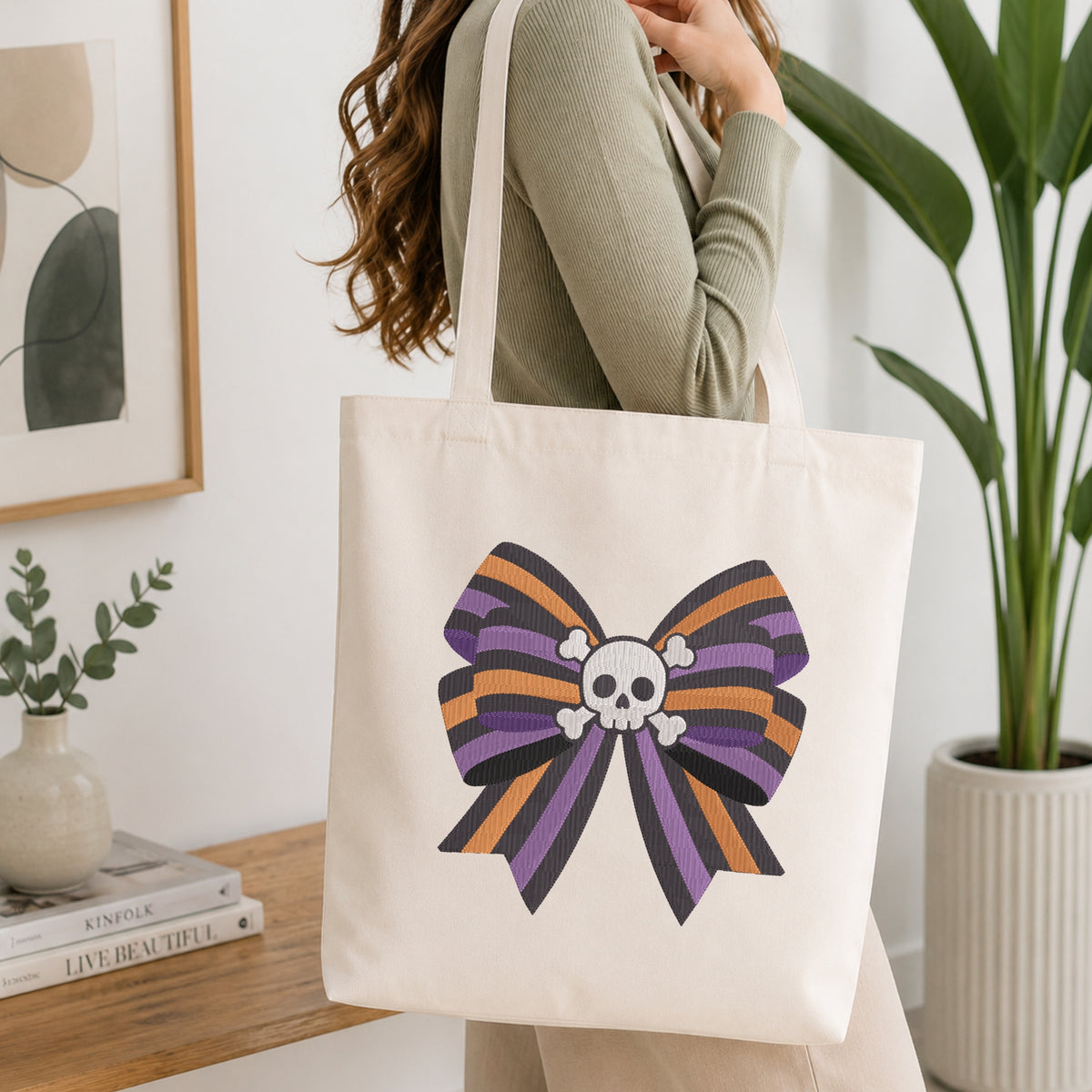 A person wearing a green long-sleeve top and carrying a natural canvas tote bag printed with the striped skull bow machine embroidery graphic.