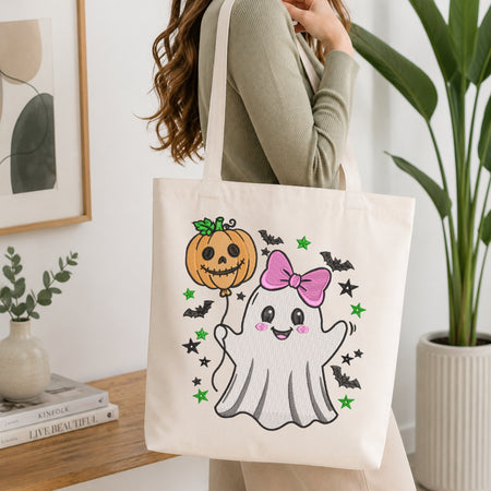 A woman wearing a sage green ribbed long-sleeve top carries a cream canvas tote bag decorated with a cute Halloween graphic of a smiling ghost with a pink bow, a jack-o'-lantern, bats, and stars.
