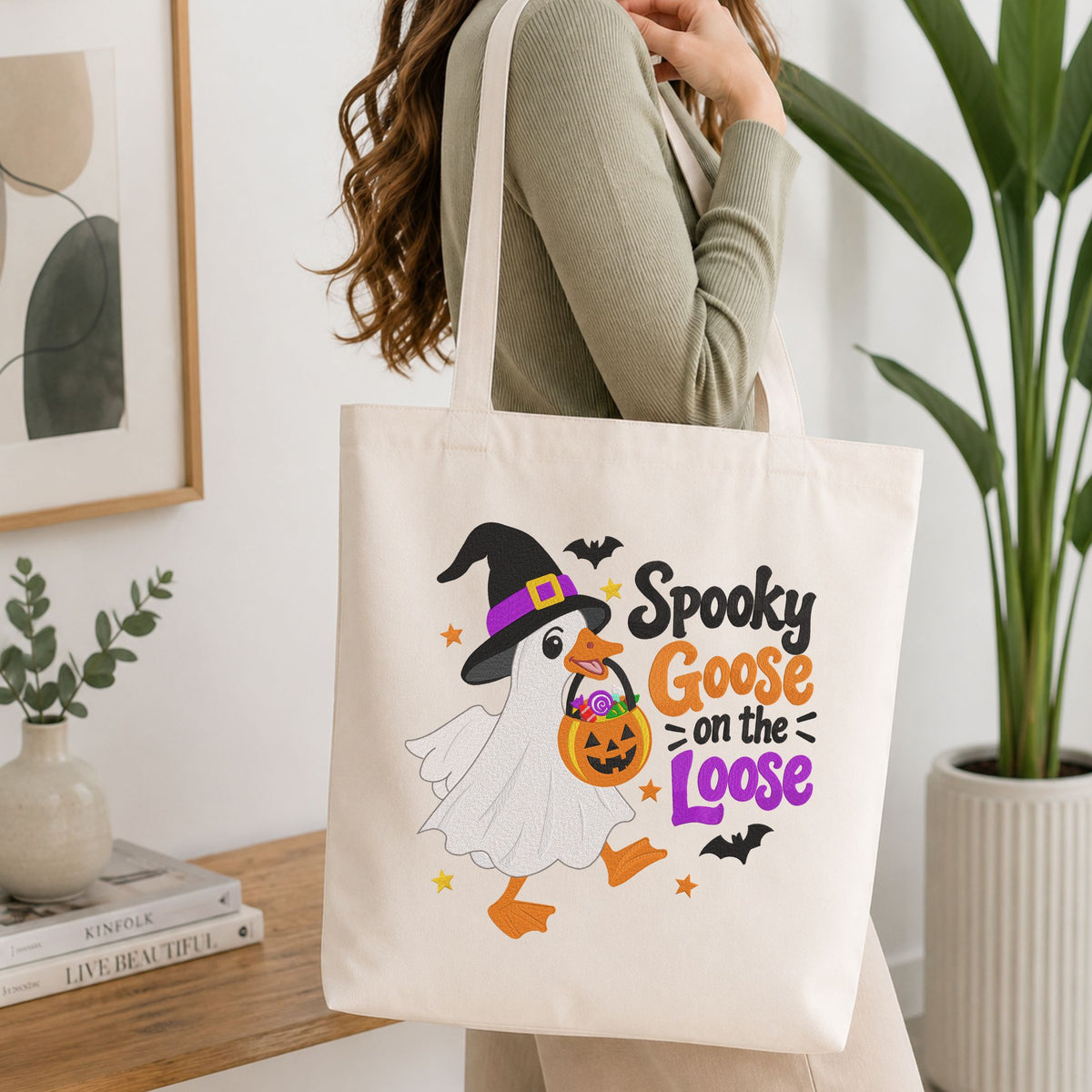Woman carrying a white natural canvas tote bag featuring a "Spooky Goose on the Loose" machine embroidery design in a bright, modern interior setting   