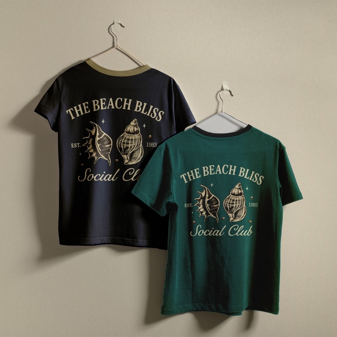 Two t-shirts, one black and one dark green, hanging on a wall displaying a matching vintage style cream sea shell illustration and coastal theme text.