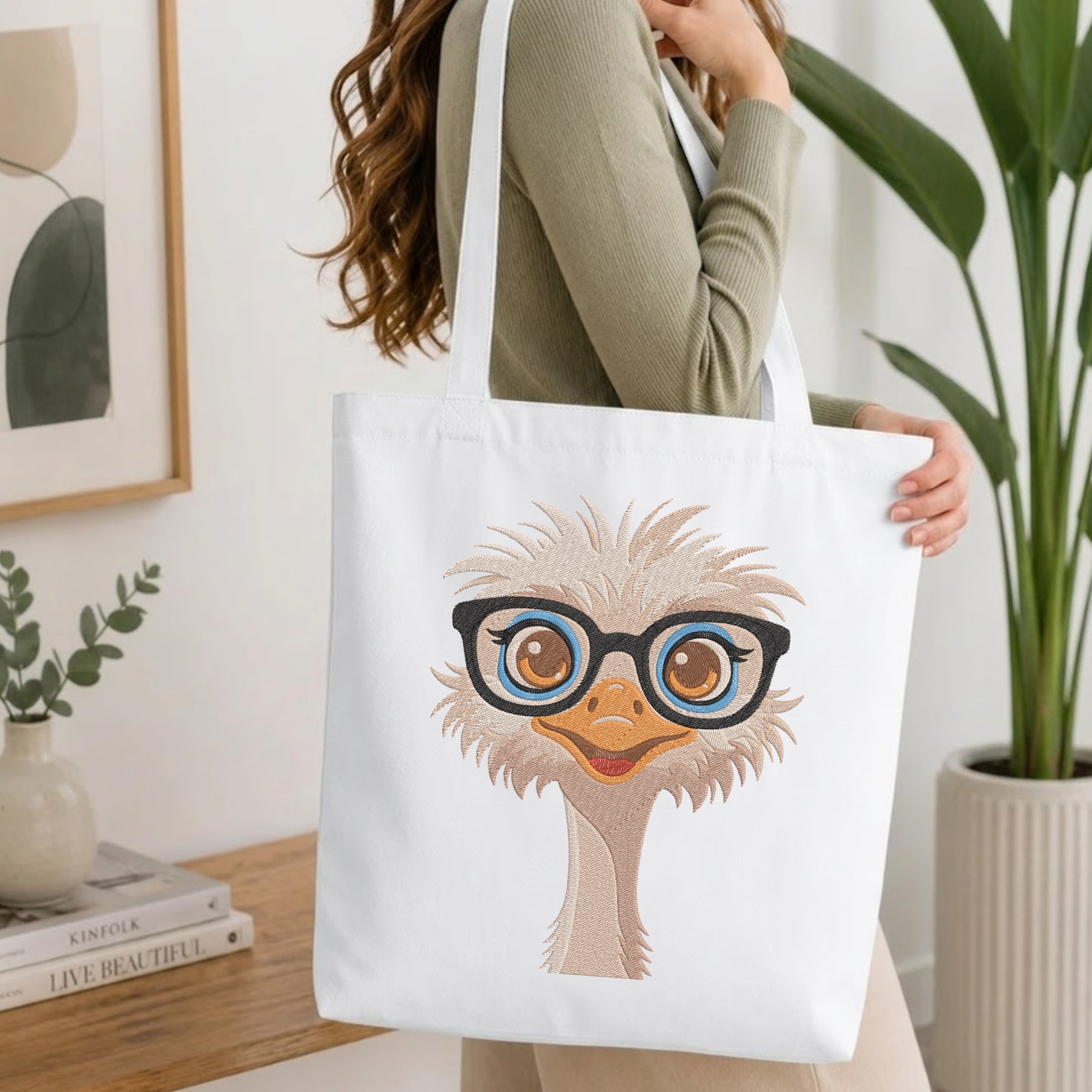 A woman wearing a sage green ribbed sweater carries a white canvas tote bag featuring a cartoon ostrich wearing black-rimmed glasses. The setting is a bright, modern room with a wooden table, books, and a potted plant.