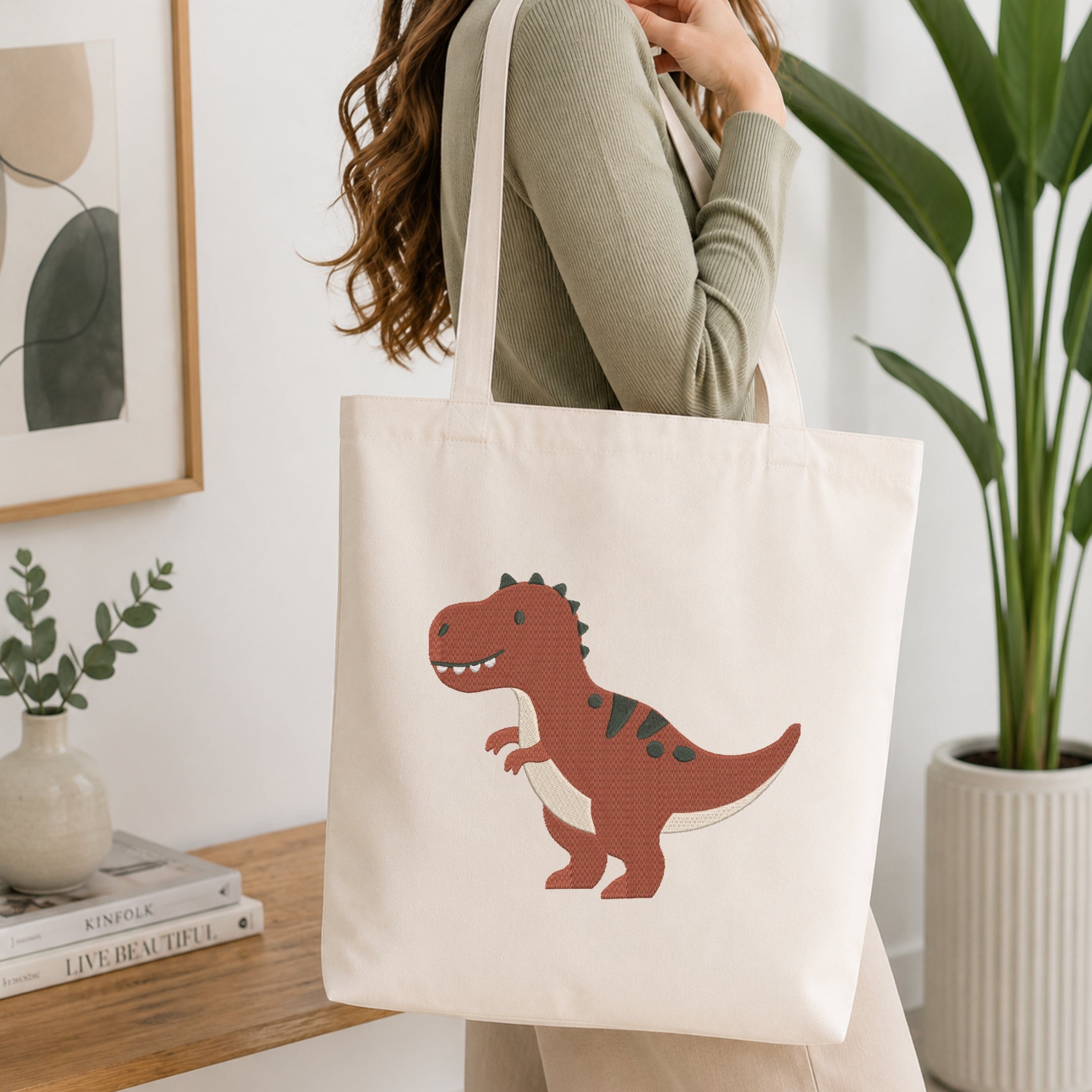 A woman wearing a sage green ribbed top carries a cream canvas tote bag featuring a cartoon T-Rex dinosaur in reddish-brown, dark green, and cream. The scene is set in a bright, modern room with a wooden table, books, a framed print, and a potted plant.