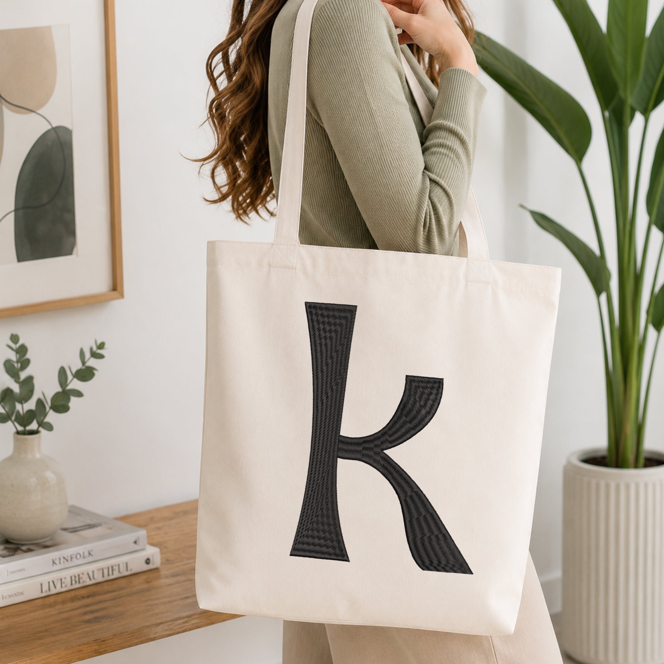 A woman wearing a sage green ribbed long-sleeve top carries a cream canvas tote bag with a large black textured letter 'K' printed on the front, set in a bright, modern room with a wooden table, books, and a potted plant.