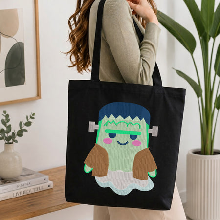 A person wearing a beige ribbed long-sleeve top carries a black canvas tote bag with a cute Frankenstein-themed monster graphic. The graphic features a light green face, brown hair, a blue hairband, and pink cheeks.