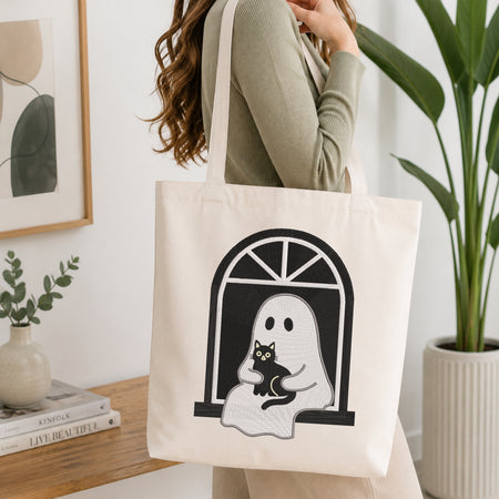 A woman wearing a sage green ribbed sweater carries a cream canvas tote bag with a black and white graphic of a ghost holding a black cat by an arched window. The scene is set in a bright, modern room with a wooden table, books, a potted plant, and framed wall art.