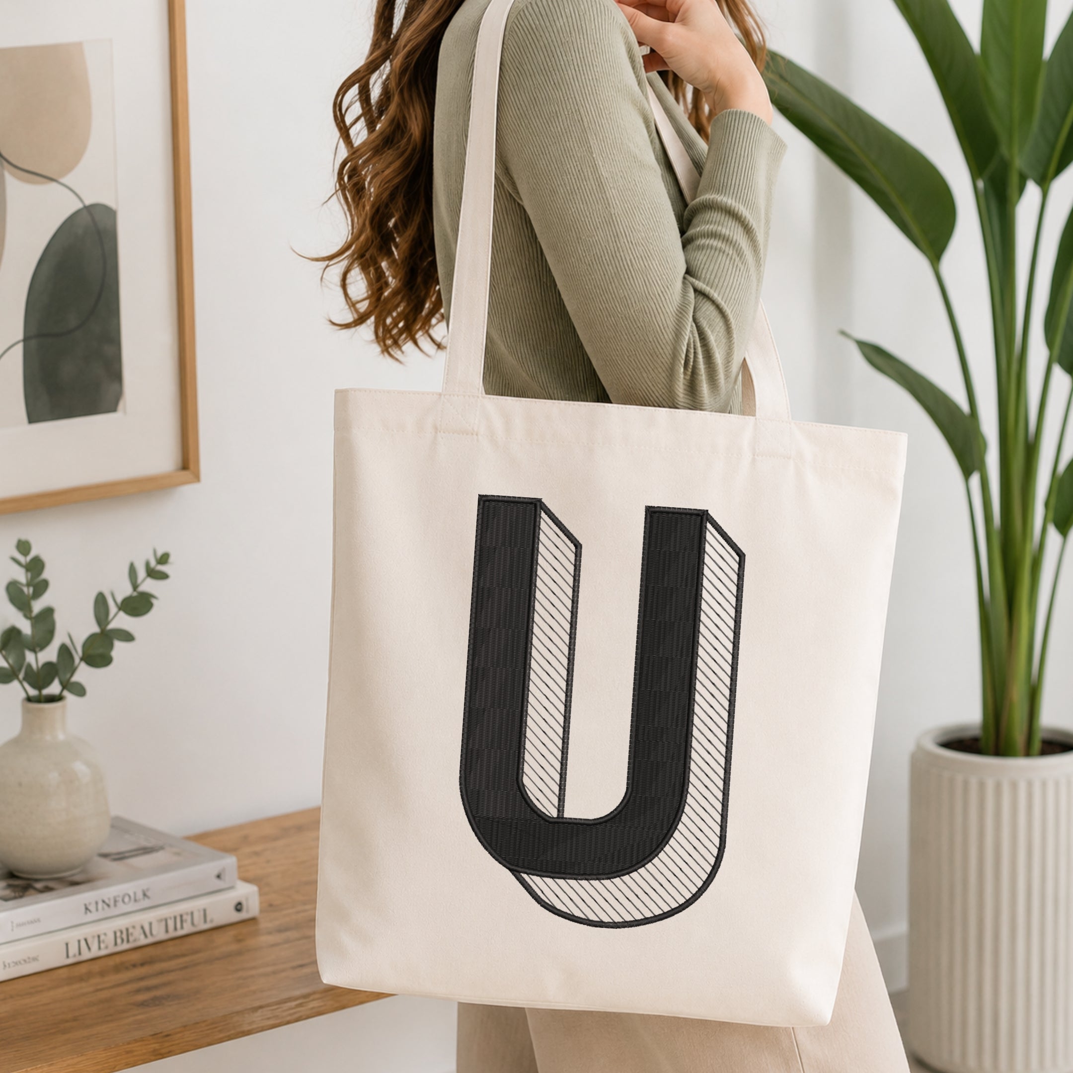 A person holding a natural canvas tote bag printed with an embroidered 3D block letter "U" design, set against a bright interior room background with green plants.