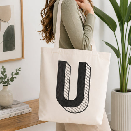 A person holding a natural canvas tote bag printed with an embroidered 3D block letter "U" design, set against a bright interior room background with green plants.