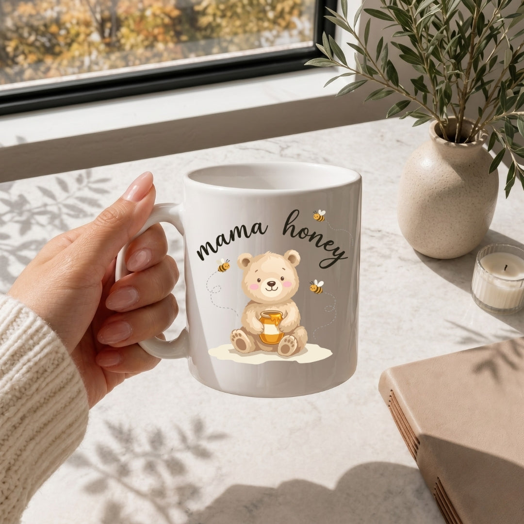White mug with 'mama honey' design held by a hand on a table with a window and plant in the background.
